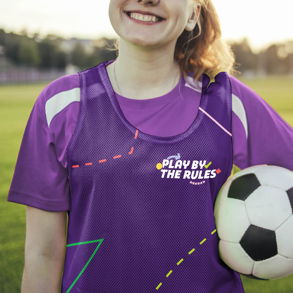 A girl smiling in a Play by the Rules sports vest. She is standing outside with a soccer ball